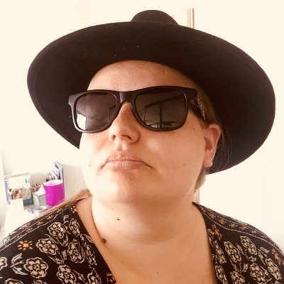 Author photo of Bex Hainsworth facing left, 3/4 wearing big dark ray-ban type sunglasses wearing a big black hat.