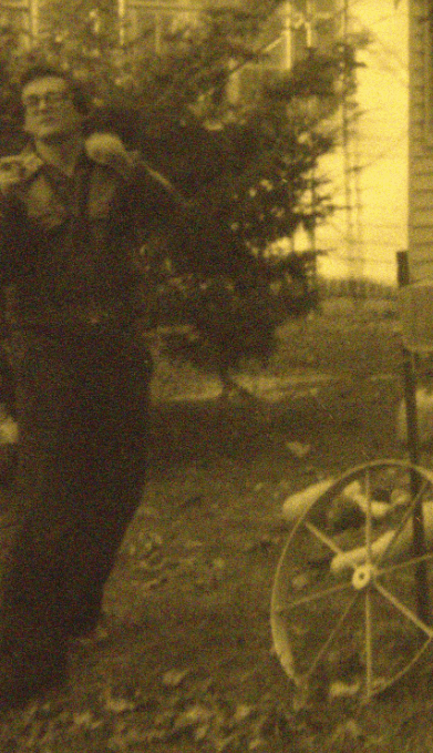 Author photo of Ernie Brill by Robert Frede Kenter -- yellow sepia - Ernie standing in a yard leather jacket, black pants, raising his hand to his face, standing in front of an old wooden house, with a wheel spoke ornament at his right, somewhere upstate NY.