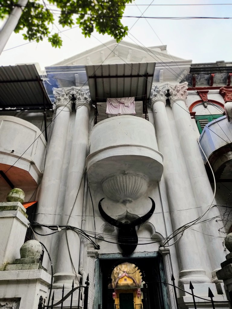 An old colonial style bank building with cornices seemingly reconstituted as a multi-purpose shrine, a close up of the facade with its black steer sculpture and jewel-crown yellow-gold entrance below the crumbling front, the hints of gates etc.