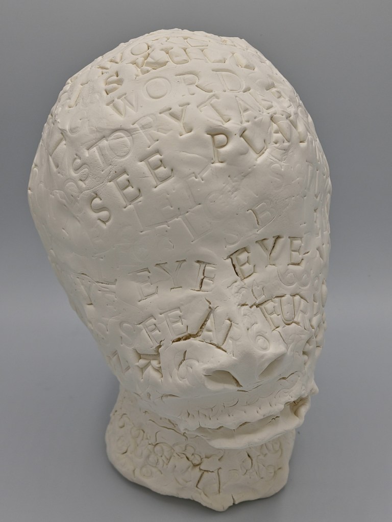 Clay Head #1 Front angled as if head is questioning something. Rough, raw sculpture with sense of movement and scattered text
 gouged stamped words: STORY SEE PLAY EYE  n wave patterns over the clay head.